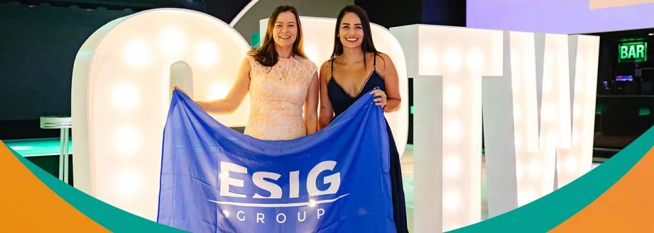 Duas mulheres segurando a bandeira da ESIG Group em um evento, com letras iluminadas ao fundo.