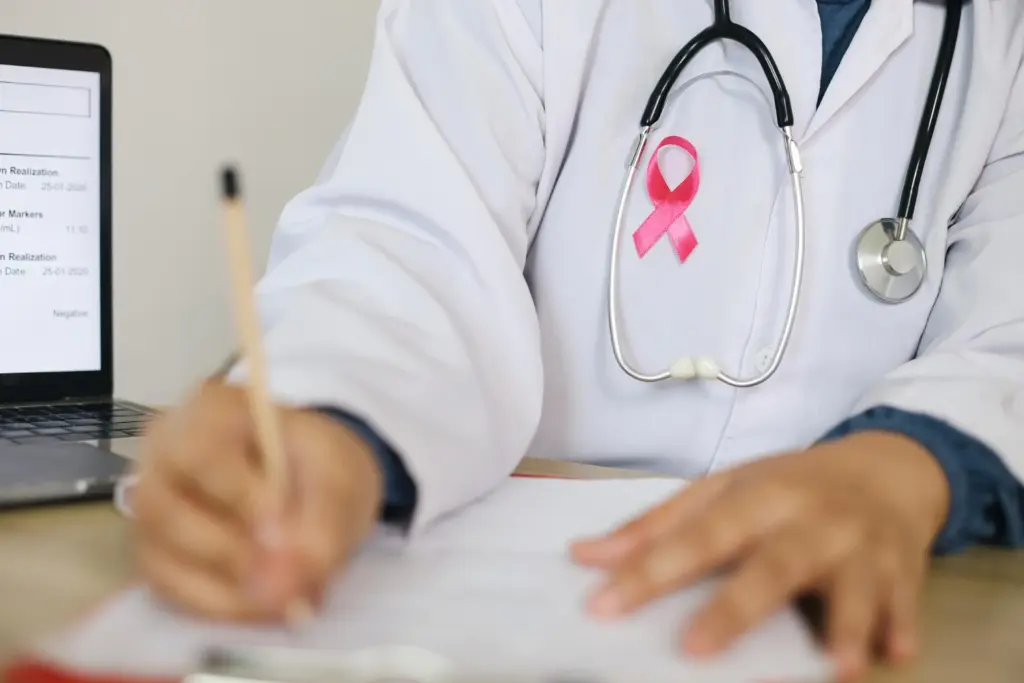 Médico com laço rosa no peito, símbolo de Outubro Rosa, ao lado de um computador e escrevendo em um papel, promovendo a conscientização sobre o câncer de mama.