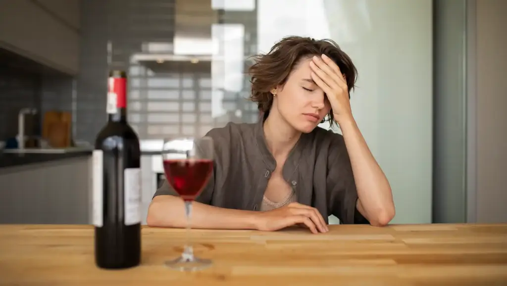 Mulher com expressão de cansaço ou dor após consumo de álcool, ao lado de uma garrafa de vinho aberta e um copo cheio, ilustrando os efeitos do consumo de álcool.