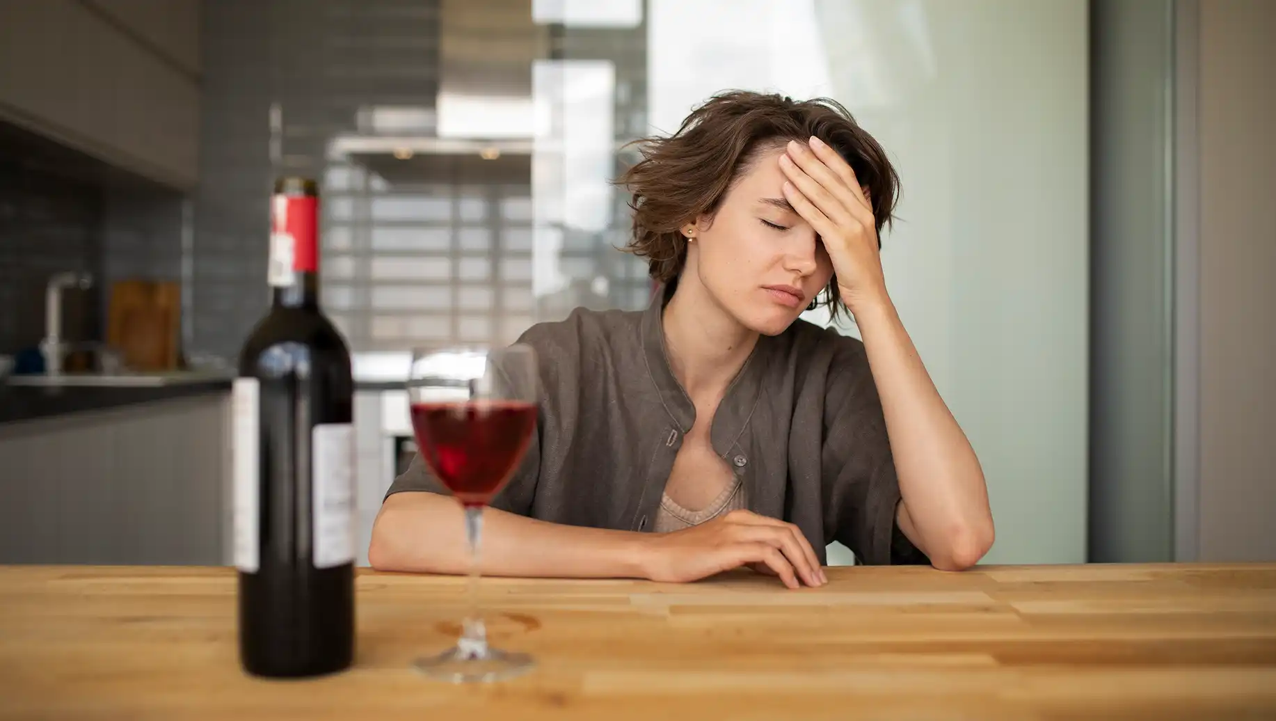 Mulher com expressão de cansaço ou dor após consumo de álcool, ao lado de uma garrafa de vinho aberta e um copo cheio, ilustrando os efeitos do consumo de álcool.