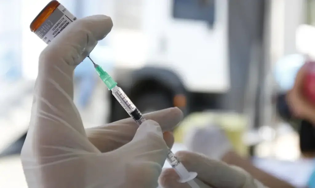 Profissional preparando vacina contra sarampo com estetoscópio em mãos e ambiente clínico ao fundo