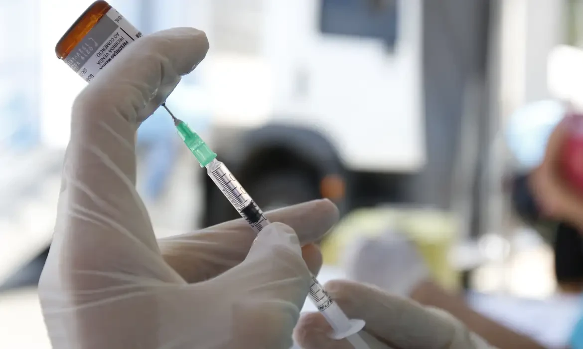 Profissional preparando vacina contra sarampo com estetoscópio em mãos e ambiente clínico ao fundo