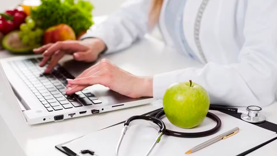 Profissional de saúde usando laptop em consultório com maçã verde, estetoscópio e frutas ao fundo, representando bem-estar e saúde.