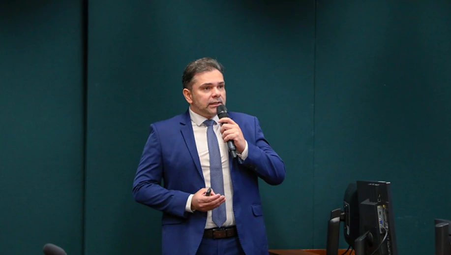 Homem em pé em um ambiente de conferência ou auditório, usando terno azul e camisa clara, segurando um microfone e falando sobre DDS para o público diante de uma mesa e equipamentos de som