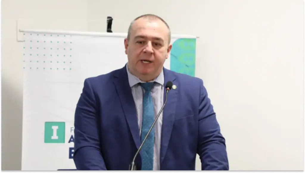 Imagem de um homem em traje formal, falando em um evento da CFM
ou conferência, com fundo de quadro branco. A imagem destaca a apresentação ou discurso de um palestrante profissional.