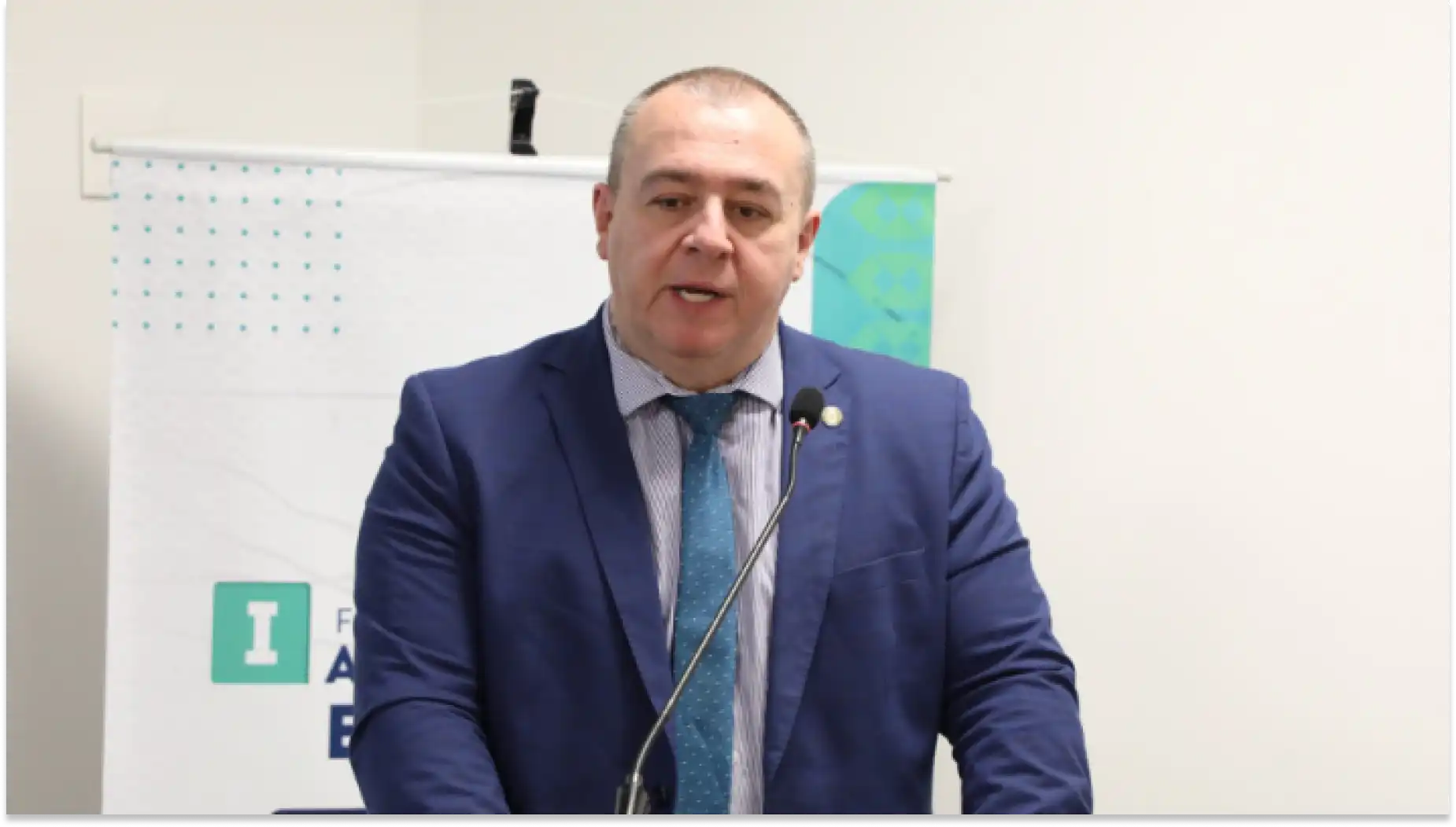 Imagem de um homem em traje formal, falando em um evento da CFM ou conferência, com fundo de quadro branco. A imagem destaca a apresentação ou discurso de um palestrante profissional.