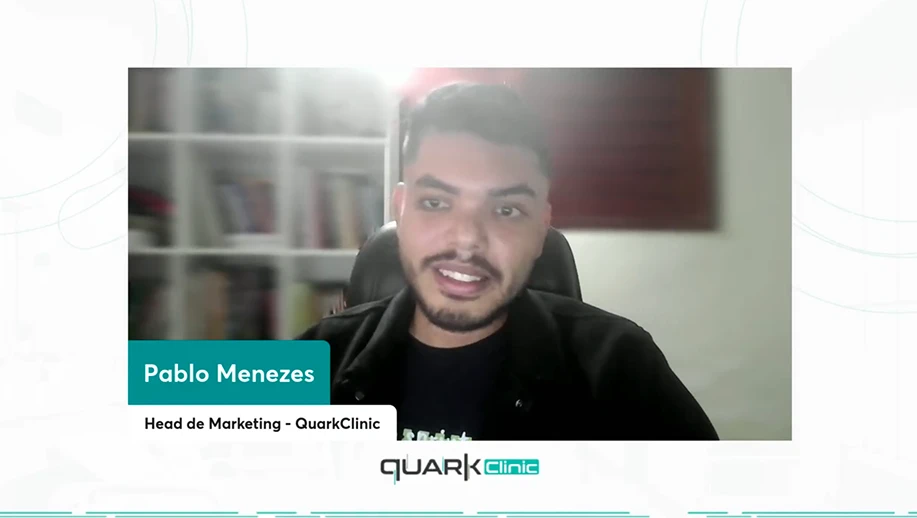 Pablo Menezes, Head de Marketing da QuarkClinic, em videoconferência, sorrindo e falando sobre marketing medico para a câmera em um fundo com estante e quadro na parede.