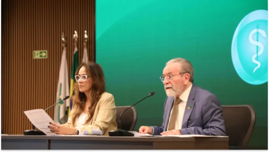 Foto de uma mesa de debate da plataforma em um auditório, com duas pessoas sentadas: uma mulher lendo anotações e um homem de terno falando ao lado de microfones. Ao fundo, painel verde com símbolo médico e uma bandeira.
