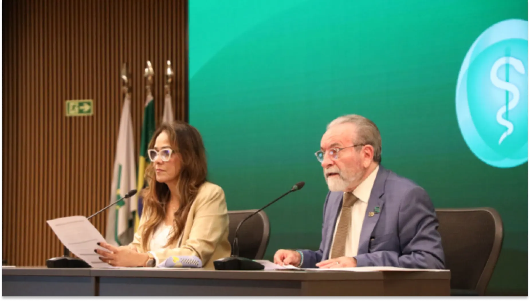 Foto de uma mesa de debate da plataforma em um auditório, com duas pessoas sentadas: uma mulher lendo anotações e um homem de terno falando ao lado de microfones. Ao fundo, painel verde com símbolo médico e uma bandeira.