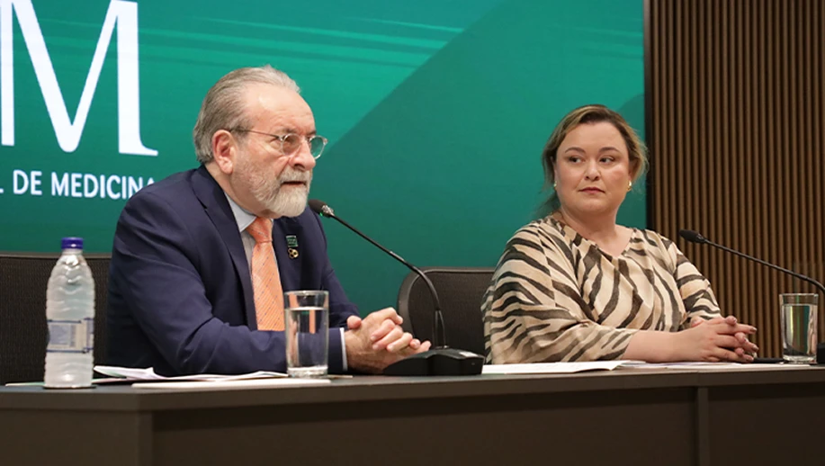 Mesa de debate sobre PMMA com dois participantes em conferência: homem idoso com barba e óculos fala em microfone, enquanto mulher ao lado observa, em painel com fundo verde e símbolos de medicina.