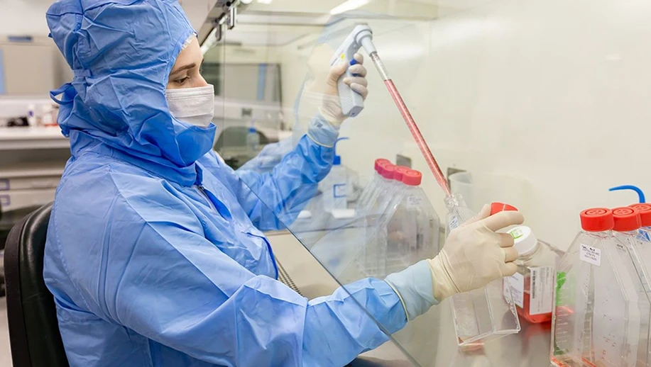 Profissional da saúde em laboratório usando capote azul e máscara, realizando procedimento com células-tronco, pipeta e tubos de ensaio com amostras em bancada.