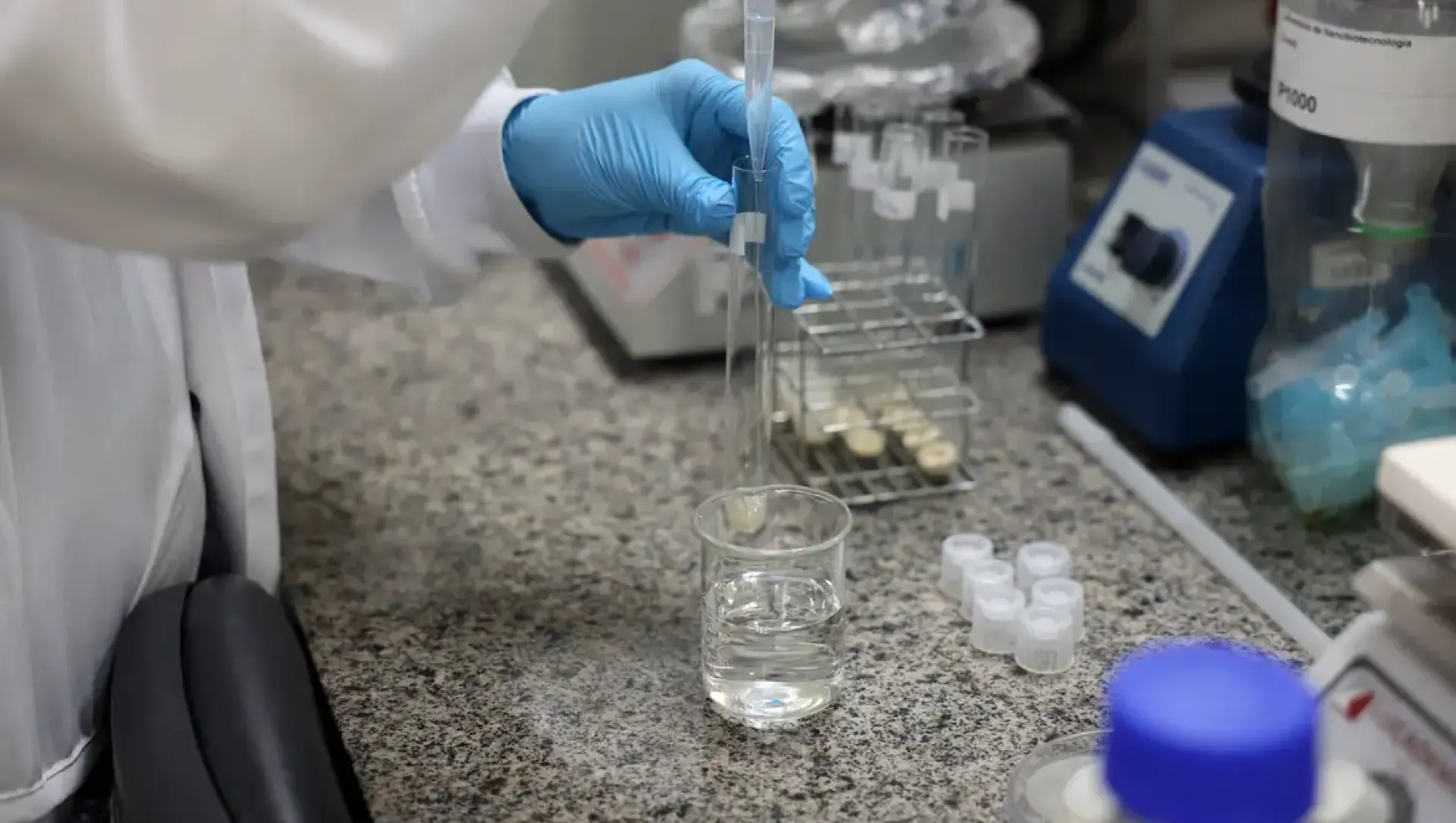 lei: Laboratório com profissional usando luvas azuis preparando amostras em tubos de ensaio e em uma bancada de granito, com frascos e recipientes ao fundo, ambiente de pesquisa