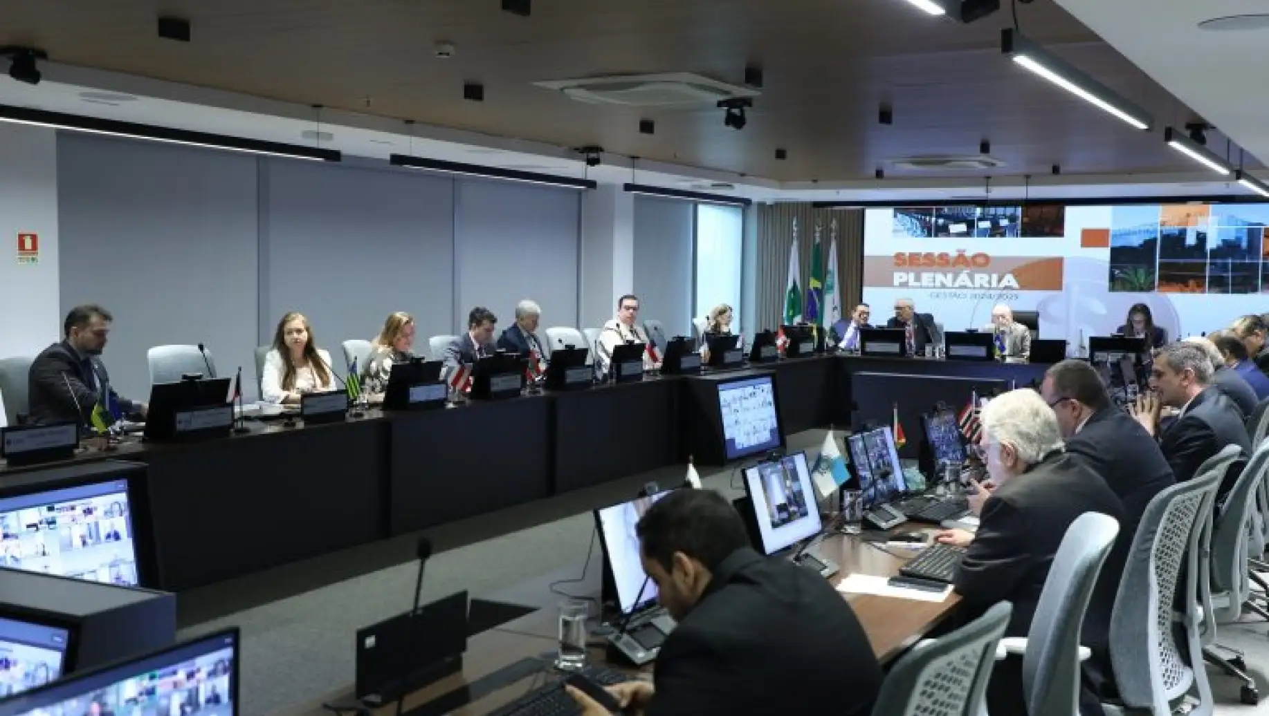 Sessão plenária em auditório com participantes sentados à mesa e painéis eletrônicos, durante reunião institucional. Regras para conflito em pauta.