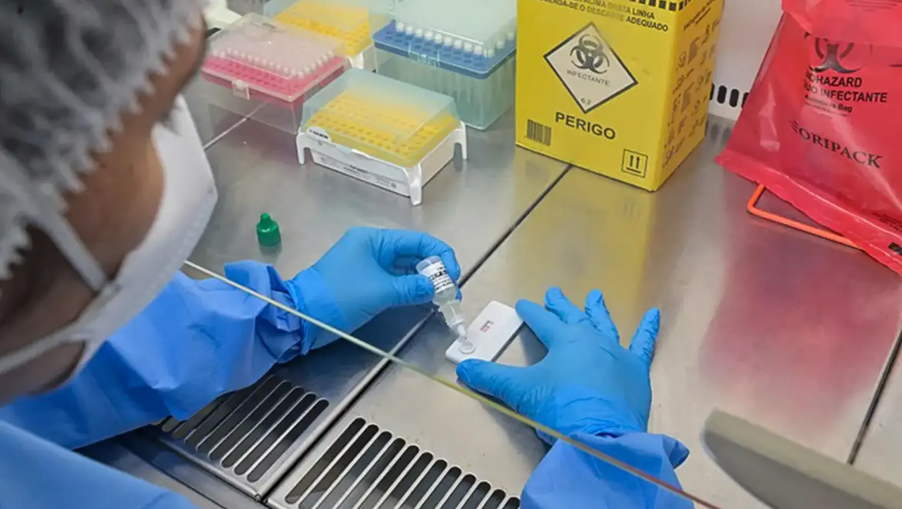 Anvisa: Profissionais com luvas azuis realizam testes em laboratório, pipetando amostras sobre uma bancada com caixas de reagentes e materiais de segurança.