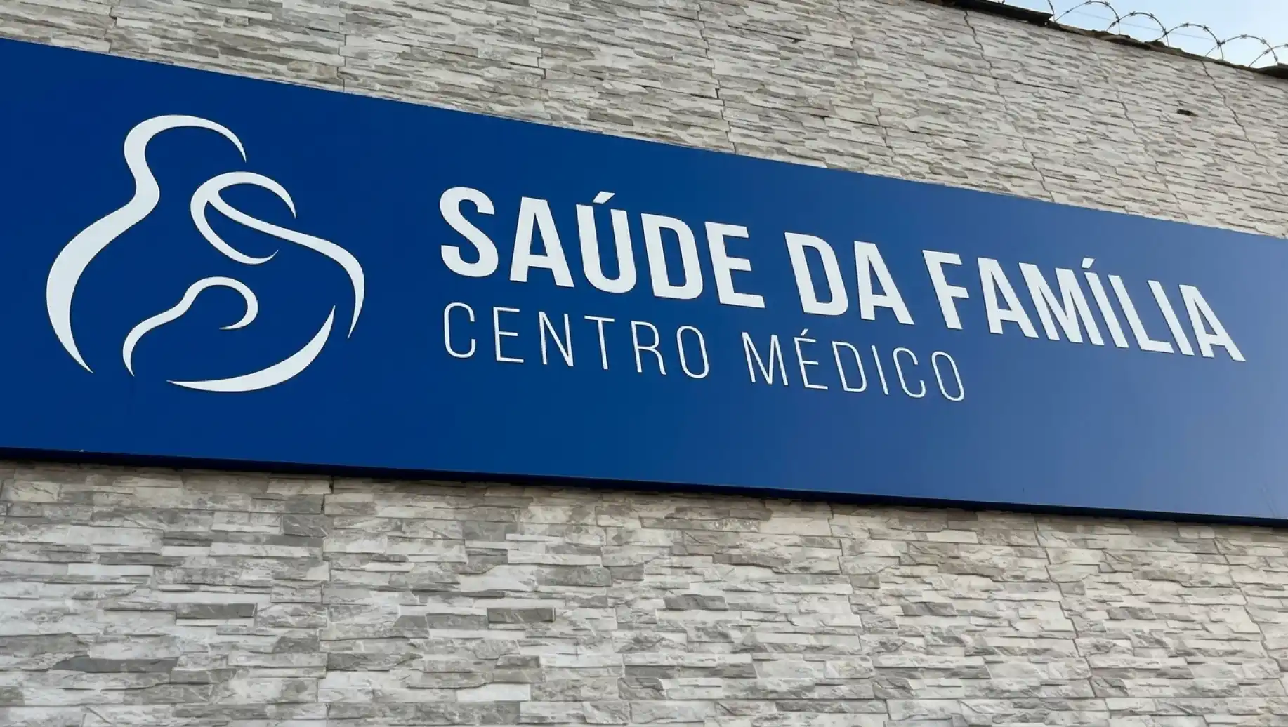 gestão: Placa externa da empresa “SAÚDE DA FAMÍLIA” com logotipo e a mensagem “CENTRO MÉDICO”, instalada em fachada de pedra, com fundo azul.