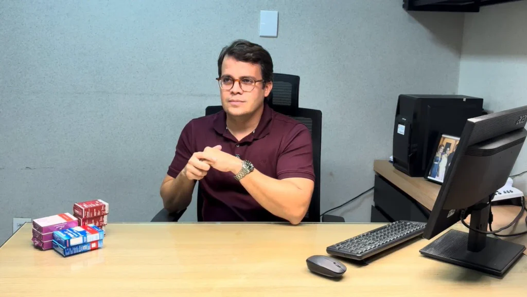 Homem em um escritório, sentado em uma mesa com computador e pilhas de livros e pastas, falando em uma apresentação sobre gestão.