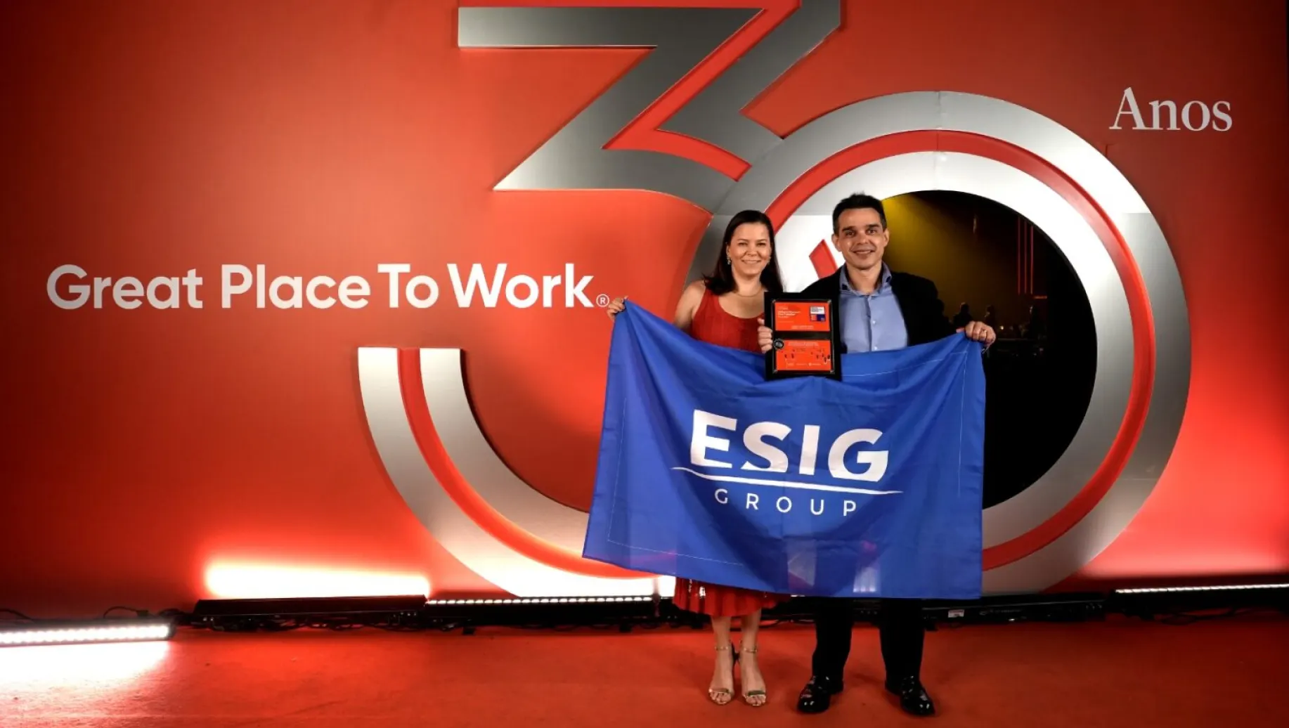 melhores empresas: Prêmio Great Place To Work em evento corporativo: casal ao lado de banner azul com o logo ESIG GROUP, segurando certificado em frente a grande logomarca 3 anos em fundo vermelho e iluminação de palco